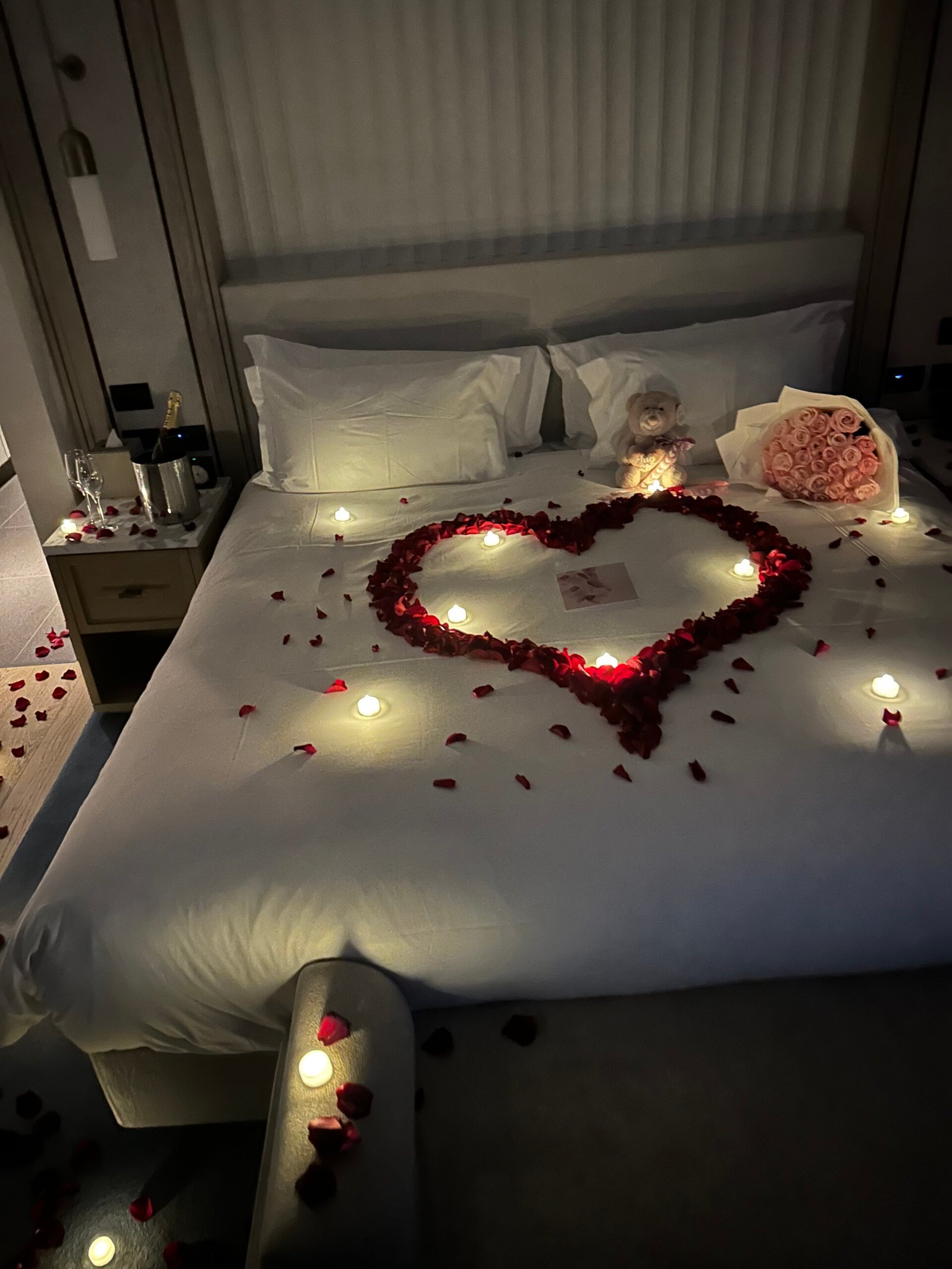 Romantic special occasion setup with rose petals, fresh flower bouquet, champagne-style bubbles and teddy bear